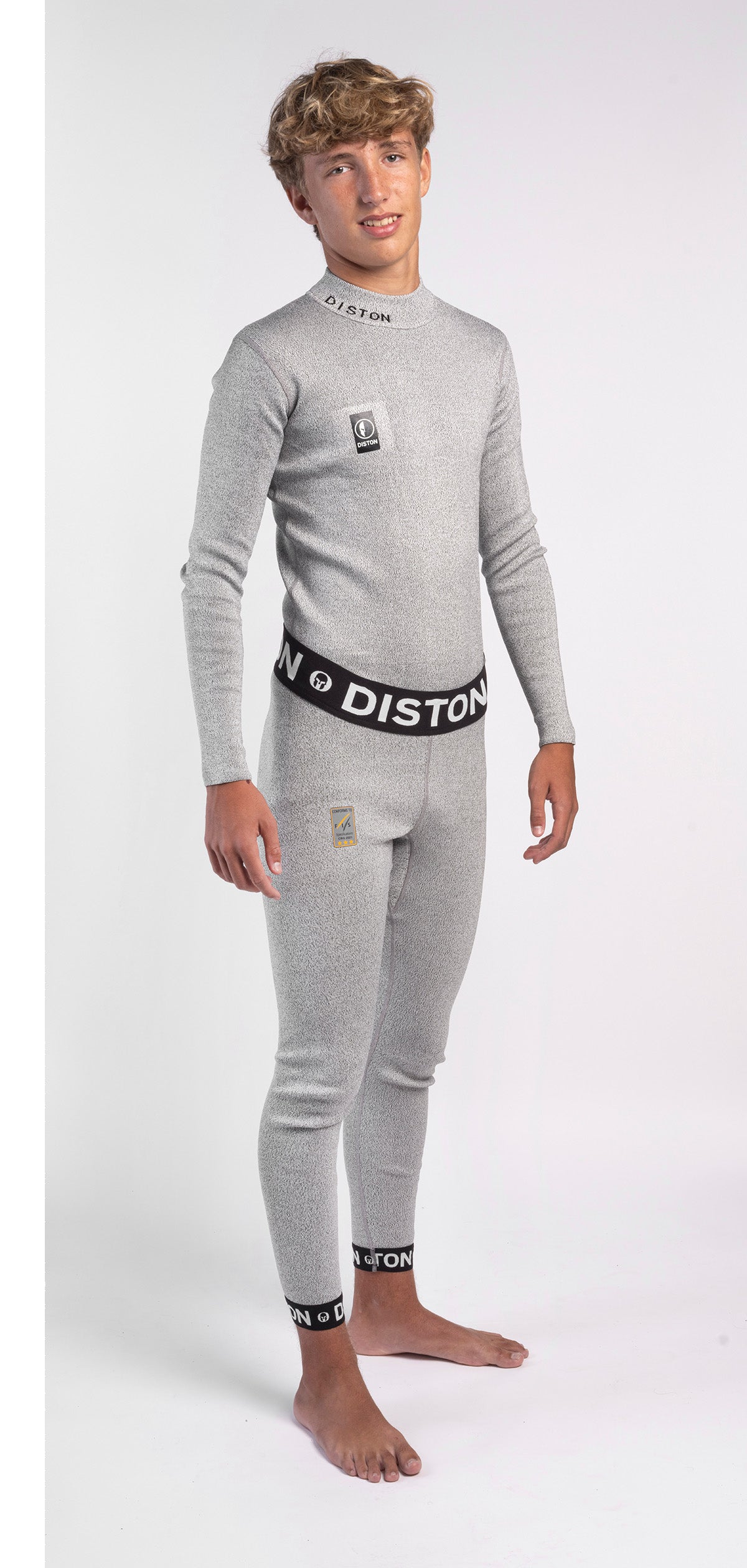 Ein Jugendlicher mit hellbraunem Haar steht barfuß und lächelt in die Kamera. Er trägt ein passgenaues Diston WC Schnittfeste Hosen Herren - 4FIS Stars in CRG - CORE / Racing Fit Set: ein hellgraues langärmeliges Oberteil und eine passende schnittfeste Unterhose mit Diston-Branding.