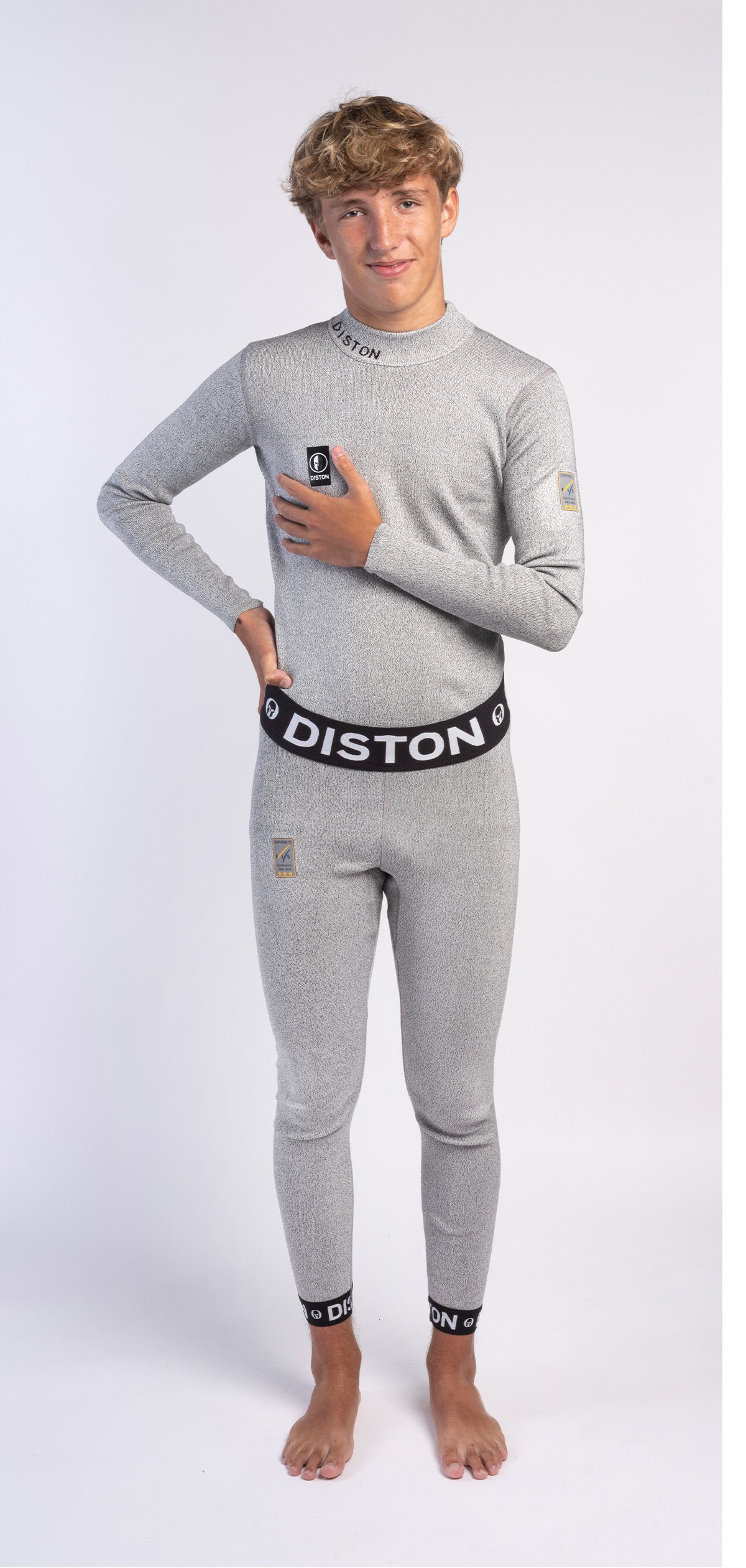 Ein junger Mensch mit hellbraunem Haar steht barfuß auf weißem Untergrund und lächelt selbstbewusst in Diston WC Schnittfeste Hosen Herren - 4FIS Stars in CRG CORE / Racing Fit-hellgraue schnittfeste Unterhose mit "DISTON" auf Bund und Bündchen, ideal für Skirennläufer.