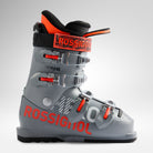 Der Rossignol HERO JR 65 - POWER GREY 2026/2027 Junior-Skischuh zeichnet sich durch rote und weiße Akzente, vier Metallschnallen, ein schwarzes Innenfutter und einen roten oberen Riemen vor einem schlichten hellen Hintergrund aus.