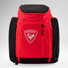 Eine rot-schwarze Rossignol HERO ATHLETES BAG mit einer Fronttasche mit Reißverschluss und dem ikonischen Rossignol-Logo, das einen Hahn auf einem Schild zeigt. Sie eignet sich ideal als Rucksack für die Skirennausrüstung und steht aufrecht vor einem hellgrauen Hintergrund.