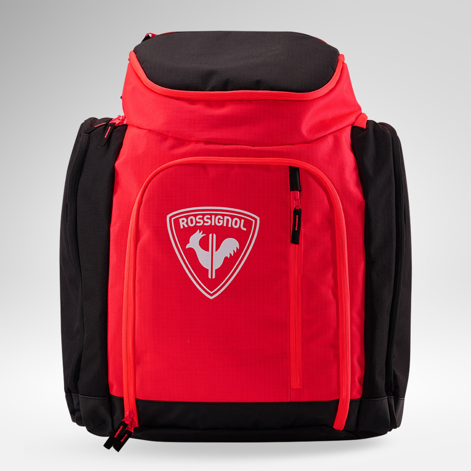 Eine rot-schwarze Rossignol HERO ATHLETES BAG mit einer Fronttasche mit Reißverschluss und dem ikonischen Rossignol-Logo, das einen Hahn auf einem Schild zeigt. Sie eignet sich ideal als Rucksack für die Skirennausrüstung und steht aufrecht vor einem hellgrauen Hintergrund.