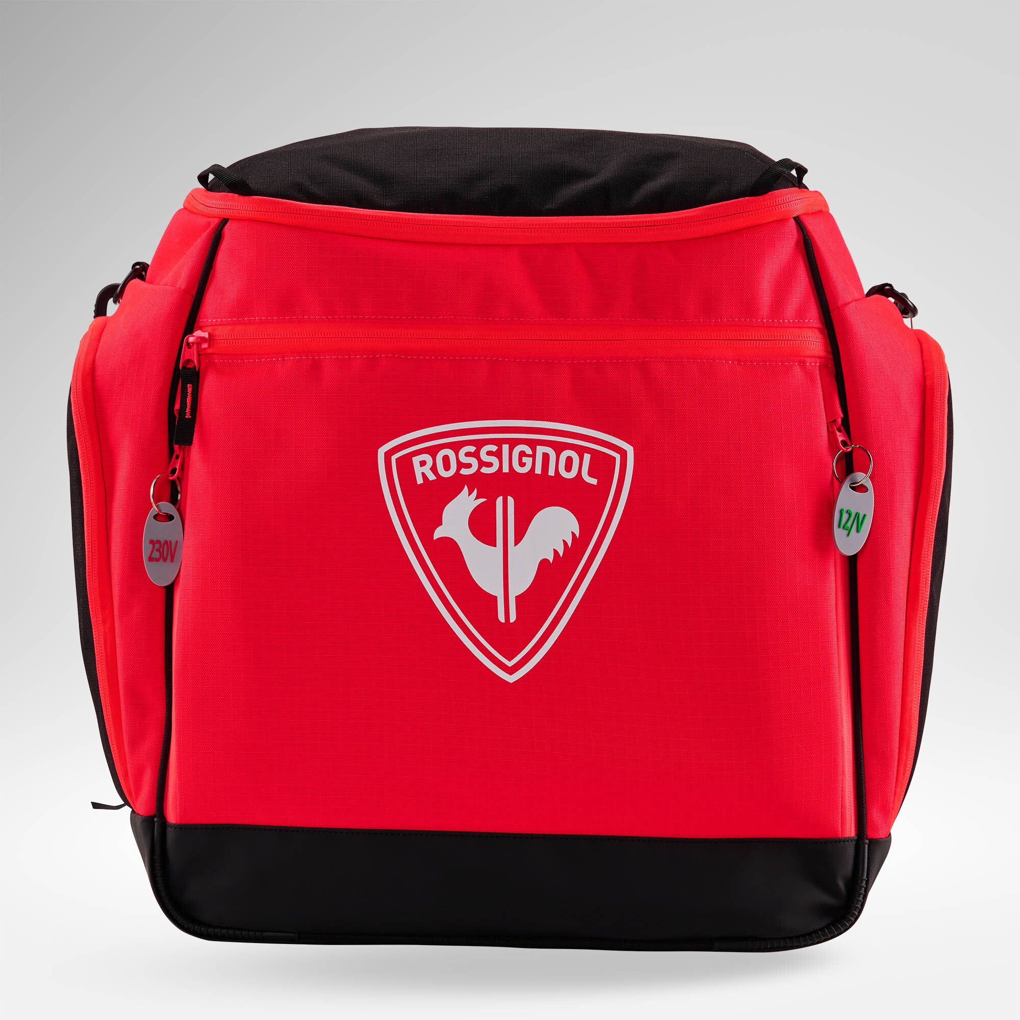 Eine leuchtend rote Rossignol HERO HEATED BAG 230V mit dem ikonischen weißen Hahn-Logo, einem robusten schwarzen Boden, praktischen Seitentaschen sowie zwei Reißverschlusslaschen mit den Aufschriften „230V“ und „12V“, sticht vor einem hellen Hintergrund mit Farbverlauf hervor – ideal für den Ski-Weltcup oder zum Warmhalten Ihrer Skiausrüstung dank integrierter Schuhheizung.