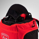 Eine Nahaufnahme des Rossignol HERO HEATED BAG 230V mit seinem markanten rot-schwarzen Design, dessen Reißverschluss geöffnet ist und der einen Skihelm sowie eine Skibrille im Inneren zeigt, verkörpert den Stil des Skiweltcups – perfekt, um Ihre Ausrüstung nach einem Tag auf der Piste warm und ordentlich aufzubewahren.