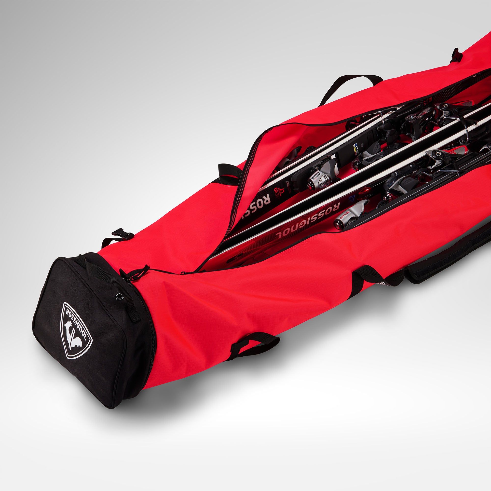 Eine rote Rossignol HERO SKI BAG 2/3P ADJUSTABLE 190/220, deren schwarze Endtasche teilweise geöffnet ist, sodass die darin befindlichen Skier und Skistöcke zu sehen sind, wird vor einem schlichten hellgrauen Hintergrund präsentiert. Diese vielseitige Tasche von Rossignol bietet reichlich Platz für Ihre Ausrüstung und ist in der Länge verstellbar (190–220 cm), was sie zur idealen Wahl für Wintersportbegeisterte macht.
