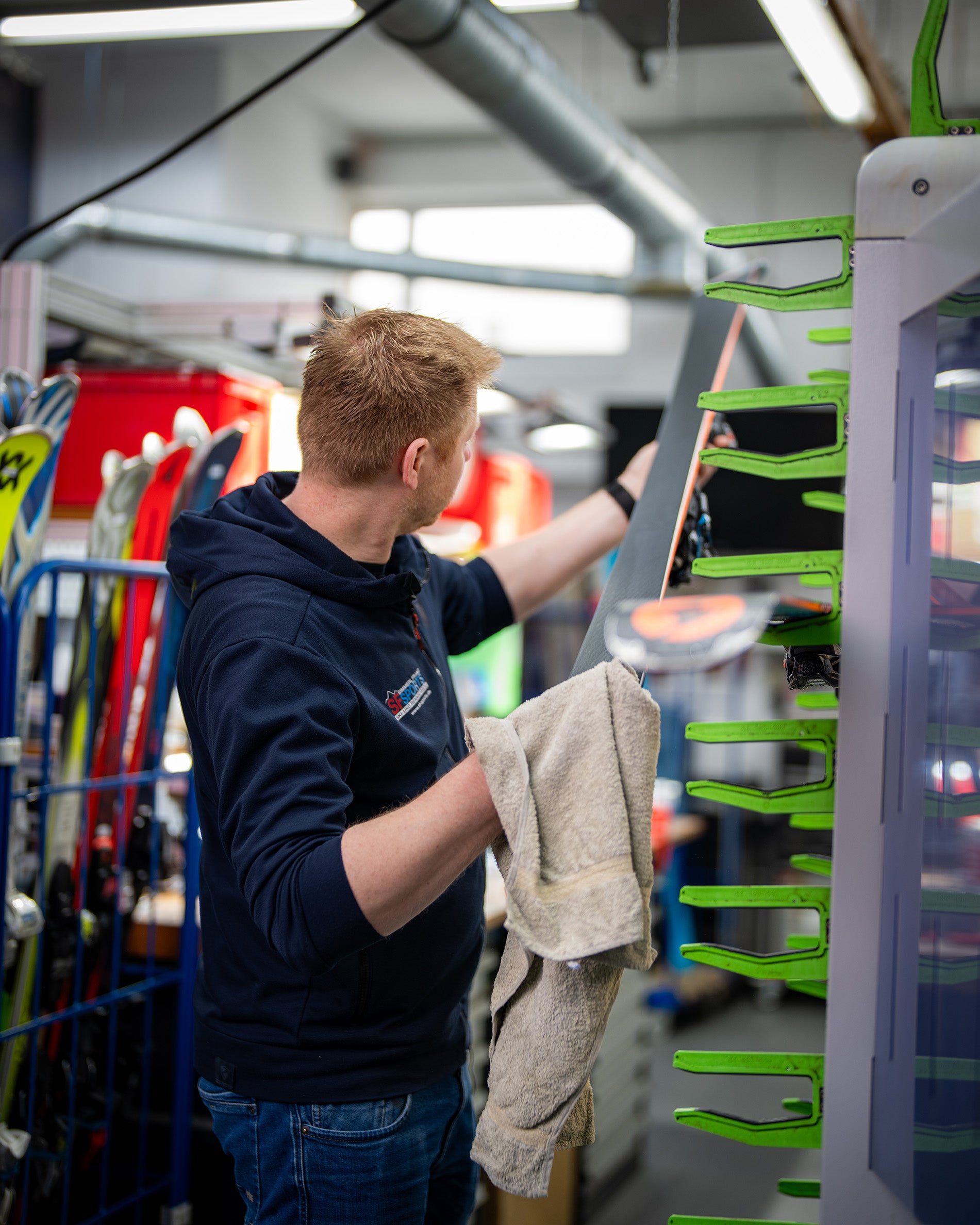 Eine Person in einem marineblauen Kapuzenpulli reinigt oder wartet Skier, hält ein Handtuch in einer Hand und arbeitet an einem Ski, der in einem grünen Gestell in einer mit Ausrüstung gefüllten Werkstatt steht.