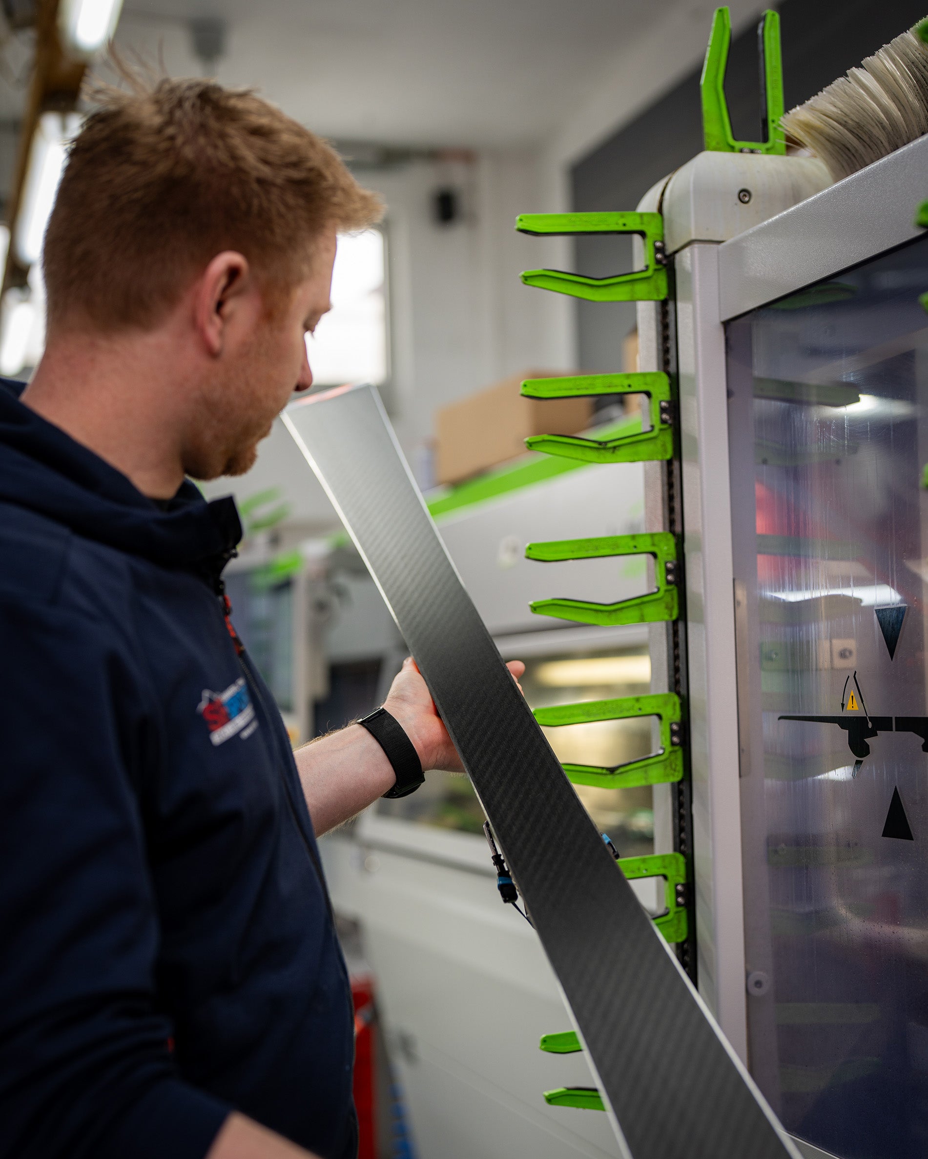 Eine Person inspiziert oder bearbeitet einen Ski in einer Werkstatt und hält den Ski in der Nähe einer Maschine mit grünen Klemmen und sichtbaren Sicherheitswarnschildern.