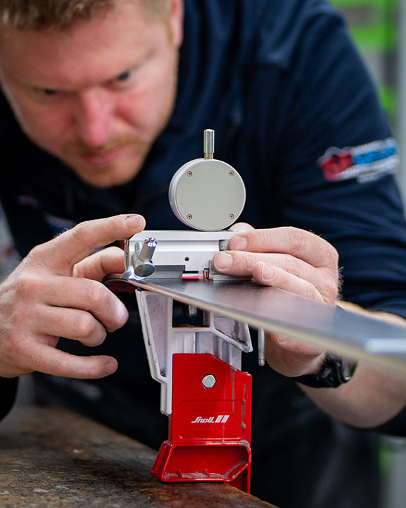 Eine Person prüft eine Skikante mit einem Messgerät und stellt den Ski, der auf einer Werkbank in einem roten Skituning-Schraubstock gehalten wird, mit großer Aufmerksamkeit ein.