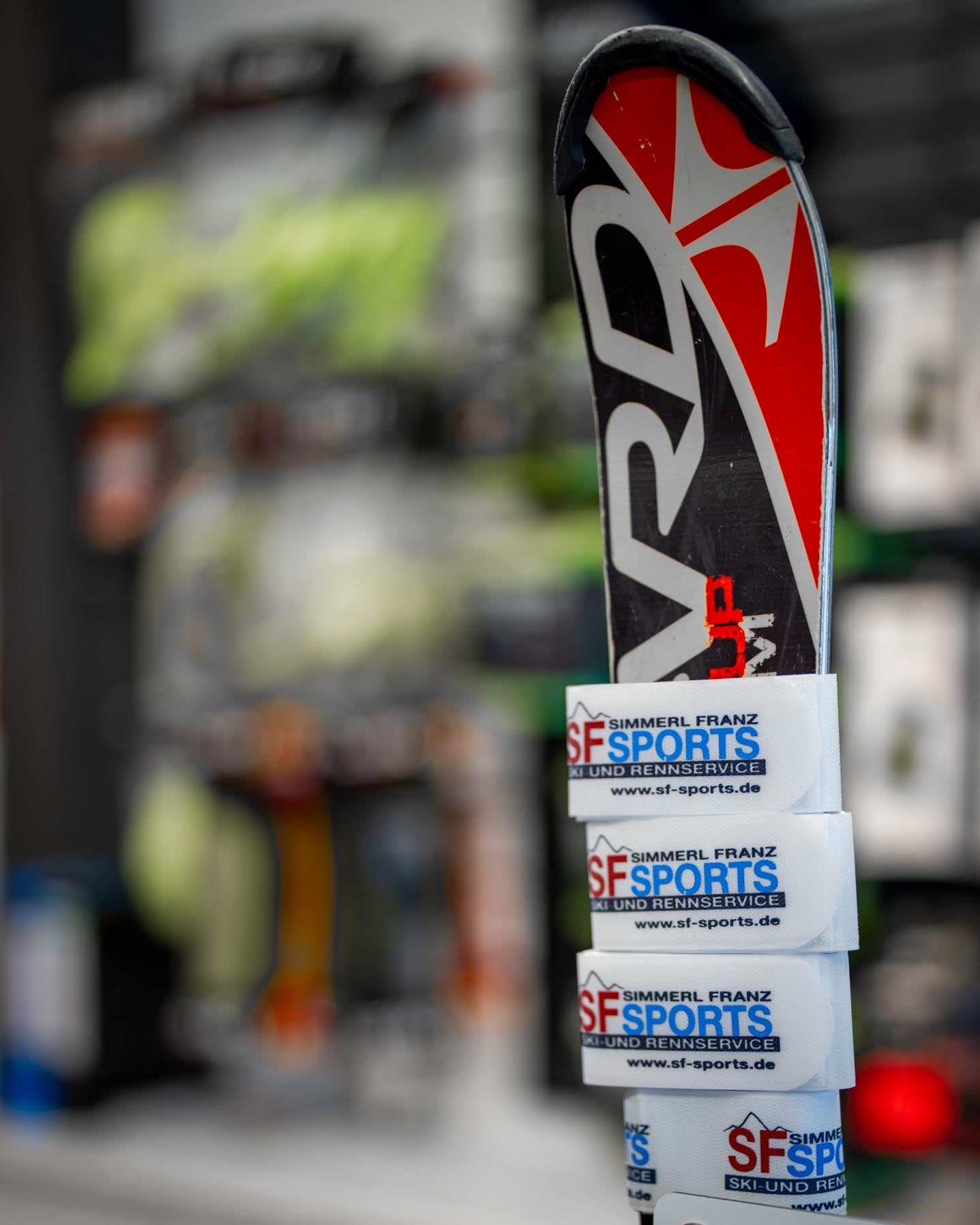 Ein Ski mit SF Sports Sticker, fotografiert im Ladengeschäft. Der Hintergrund ist verschwommen, aber die Atmosphäre im Geschäft vermittelt Professionalität und Qualität im Bereich Skisportausrüstung.
