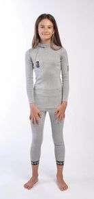 Ein junges Mädchen mit langen braunen Haaren steht barfuß auf einem weißen Hintergrund und trägt das Diston DISTON Cut Resistant Top Junior - 3FIS Stars in CRG - BASE / Performance Fit mit passenden Leggings, beide mit schwarzem Bündchentext. Sie lächelt sanft, die Hände ruhen auf ihren Oberschenkeln.
