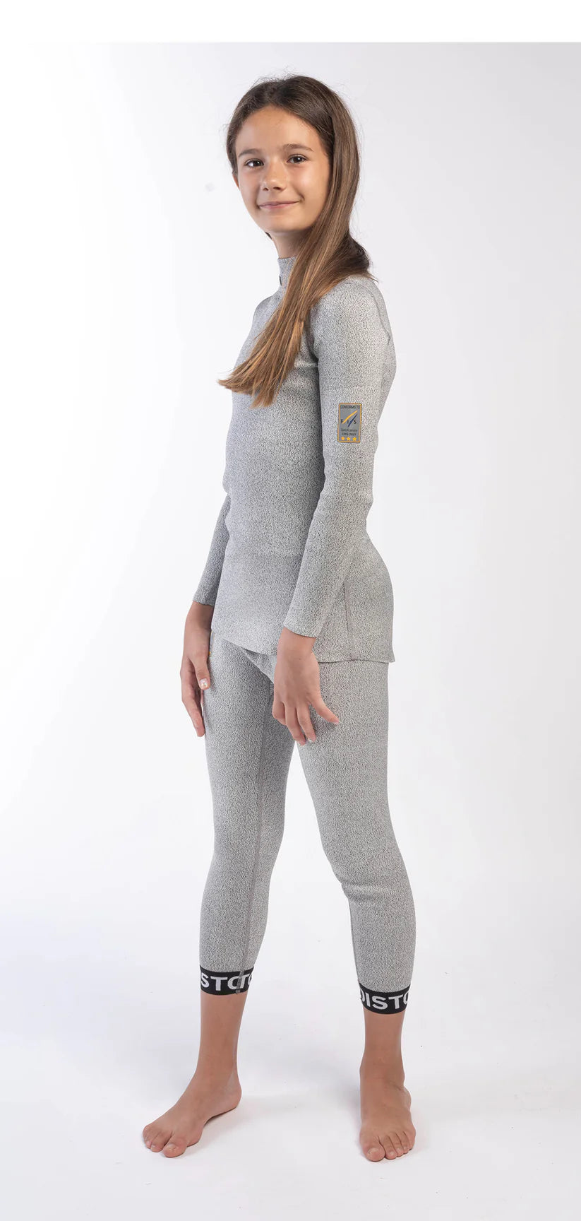 Ein Mädchen steht barfuß auf einem weißen Hintergrund und trägt das Diston DISTON Cut Resistant Top Junior - 3FIS Stars in CRG - BASE / Performance Fit-perfekt für junge Skirennfahrer. Sie schaut leicht zur Seite mit einem sanften Lächeln.