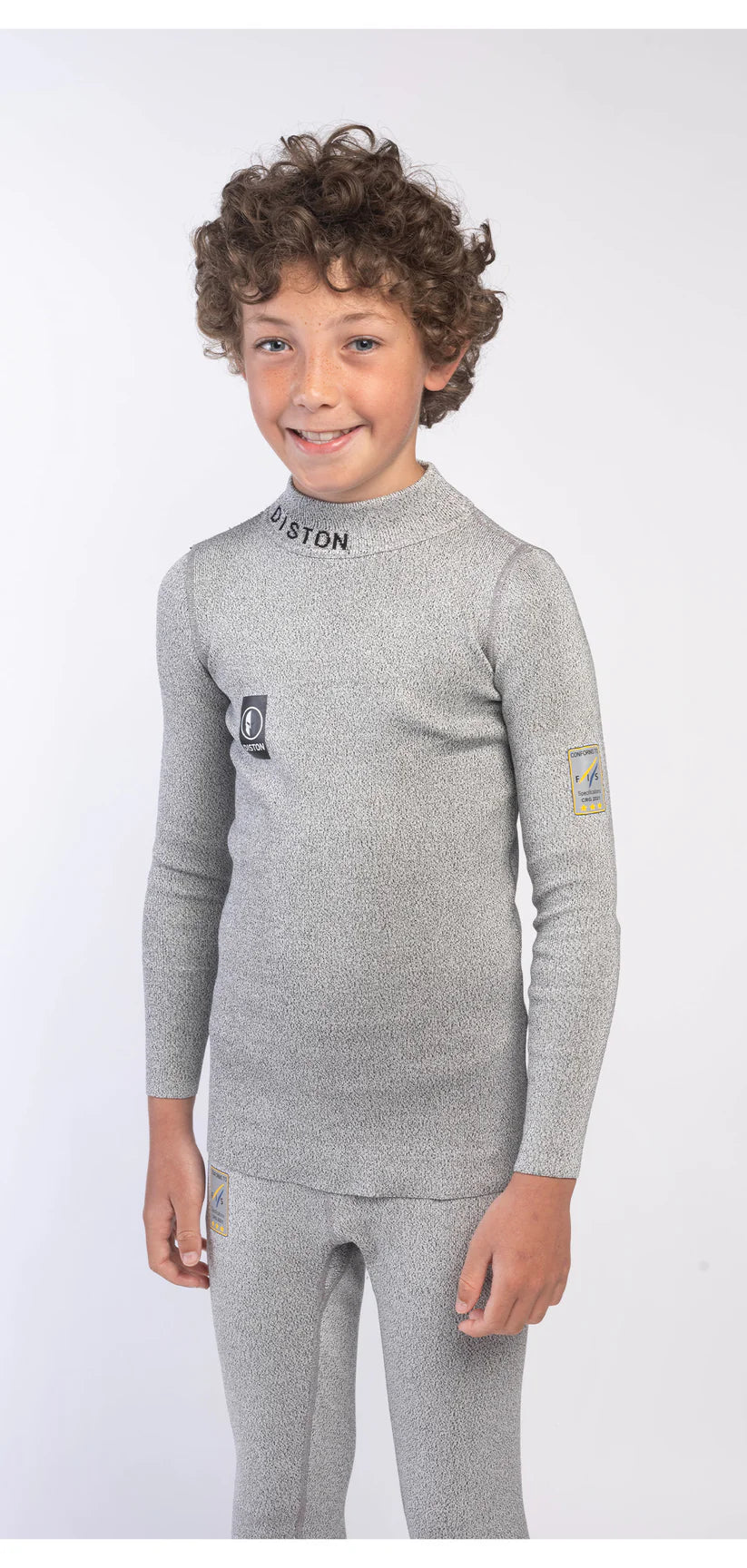 Ein kleiner Junge mit lockigem Haar lächelt, während er das Diston DISTON Cut Resistant Top Junior - 4FIS Stars in CRG - CORE / Racing Fit trägt, ein hellgraues, sportliches Top mit schwarzen und gelben Patches, ideal für Rennläufer, vor einem weißen Hintergrund.