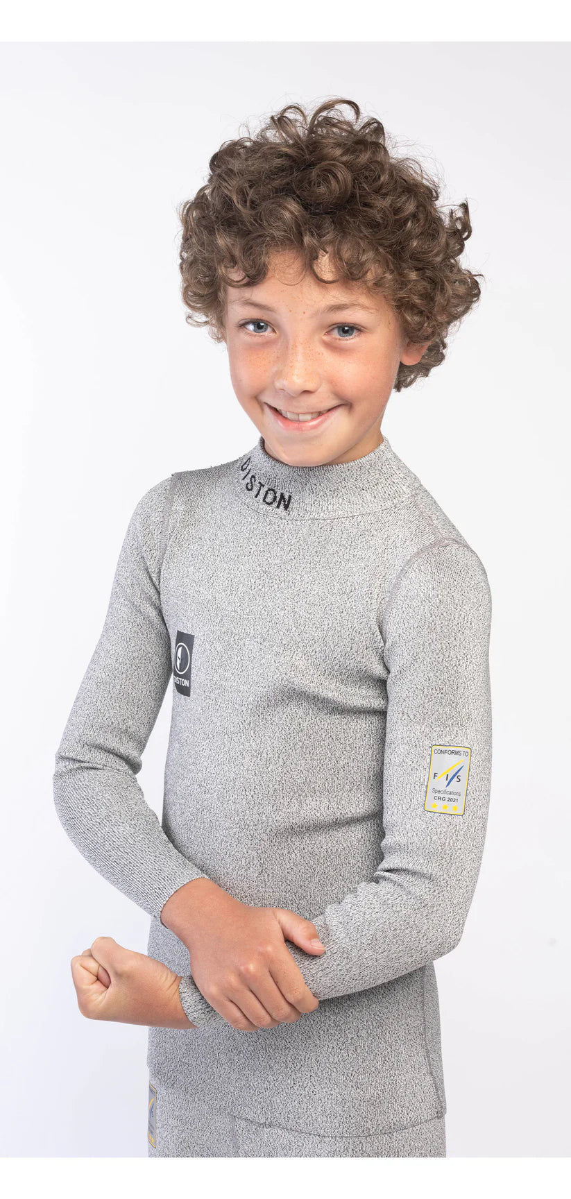 Ein lächelndes Kind mit lockigem Haar trägt das Diston DISTON Cut Resistant Top Junior - 4FIS Stars in CRG - CORE / Racing Fit, grau und langärmelig, und posiert selbstbewusst - der ideale Schutz für junge Skirennfahrer.