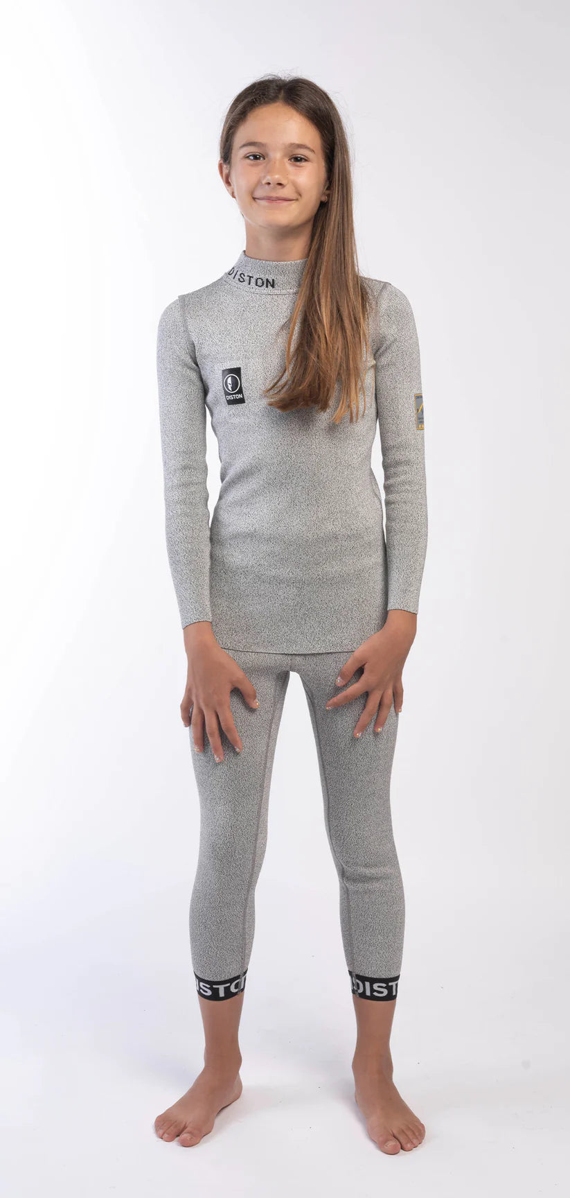 Ein junges Mädchen mit langen braunen Haaren steht barfuß vor einem schlichten weißen Hintergrund und trägt das Diston DISTON Cut Resistant Top Junior - 3FIS Stars in CRG - BASE / Performance Fit mit passender Leggings und schwarzen und weißen Markenakzenten.