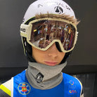 Eine Person in blau-weißer Sportjacke mit Aufnähern trägt einen Diston DISTON Cut Resistant Neck Guard (4 FIS Sterne, CRG - CORE / Racing Fit), einen Skihelm und eine Skibrille, die den hell erleuchteten Raum reflektiert - ideal für den Skirennlauf.