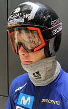 Eine Person trägt einen schwarzen Skihelm, eine orange Skibrille und den Diston DISTON Cut Resistant Neck Guard - 4 FIS Sterne in CRG - CORE / Racing Fit. Er trägt eine blaue Jacke mit Movistar- und Descente-Logos - die ideale Ausrüstung für den Skirennlauf. Der Hintergrund ist schlicht und dunkel.