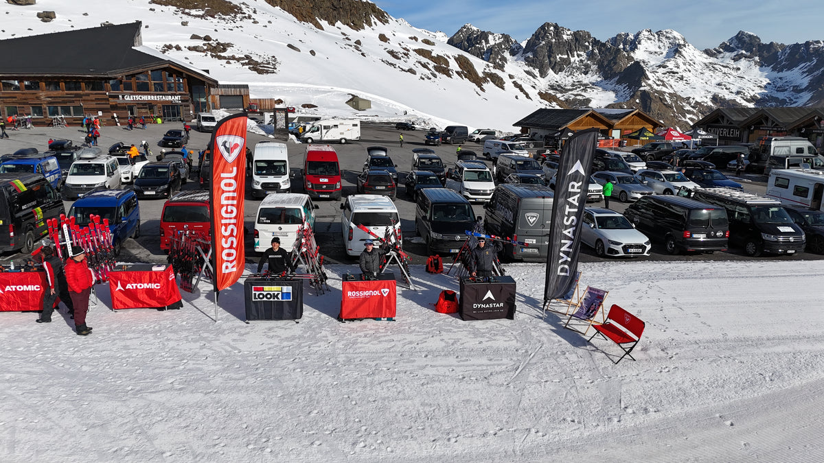 Skiausrüstungsstände mit roten Bannern, die auf dem Schnee neben einem Parkplatz voller Autos und Lieferwagen aufgebaut sind, umgeben von verschneiten Bergen und Holzhütten unter einem strahlend blauen Himmel.