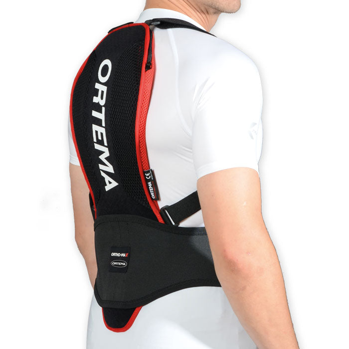 Eine Person trägt einen schwarz-roten Ortema ORTHO-MAX Light Rückenprotektor über einem weißen Hemd. Der Ortema Wirbelsäulenschutz bedeckt die Wirbelsäule vom unteren bis zum oberen Rücken und wird mit schwarzen Gurten an Taille und Schultern befestigt.